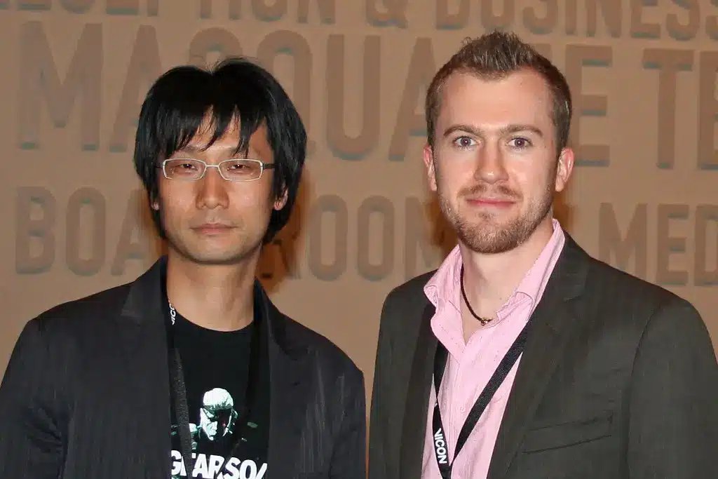 Hideo Kojima mengenakan kacamata dan blazer hitam, berdiri di samping seorang pria lain.
