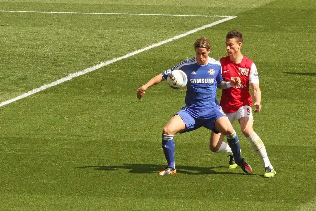 Fernando Torres dengan seragam biru Chelsea sedang mengontrol bola dengan Laurent Koscielny di belakangnya.